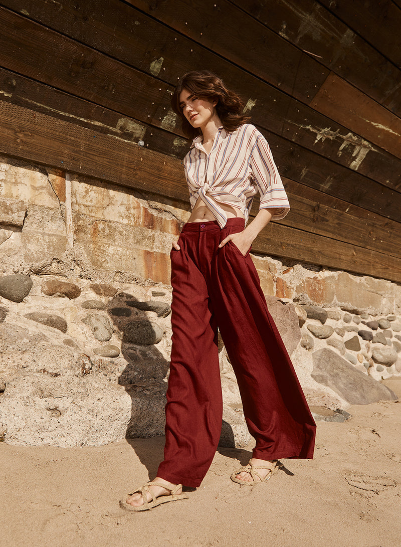 Linen Wide Leg Trouser in Bordeaux-model is walking on sand wearing a long sleeve stripped shirt and red pants